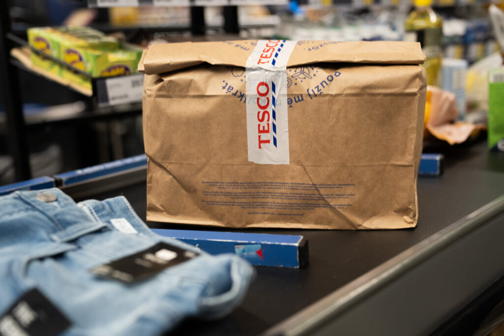 DSC03980 - Tradeinfo.cz - ForexMag.cz Brown paper package sealed with branded tape on a store checkout belt, with blurred supermarket shelves in the background.