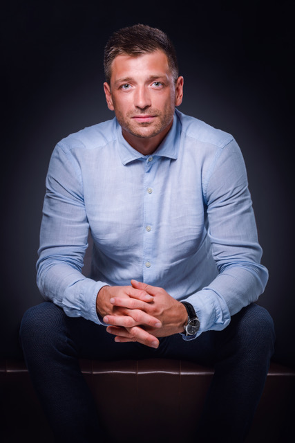 psvoren-foto - Tradeinfo.cz Man in a light blue button-down shirt sits on a bench with hands clasped, looking at the camera against a dark background.