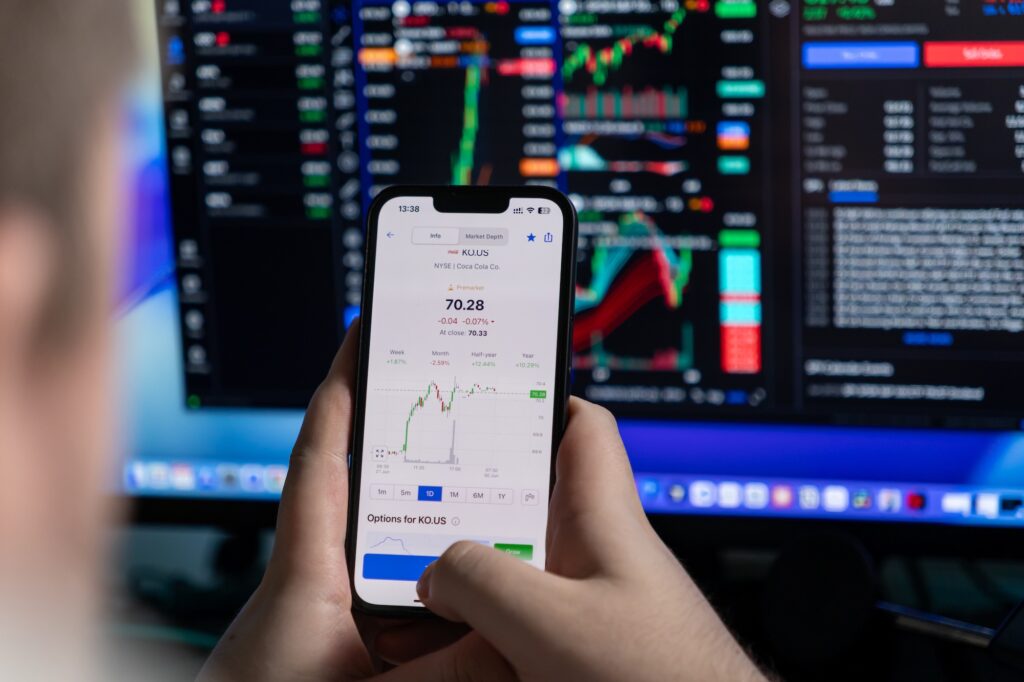 Investování - Tradeinfo.cz Close-up of a hand holding a smartphone showing Coca-Cola (KO.US) stock chart and price, with blurred trading monitors in the background.