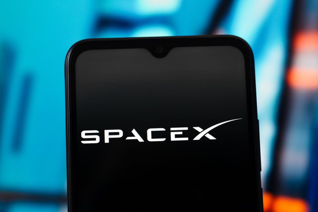Spacex - Tradeinfo.cz Close-up of a smartphone screen displaying the SpaceX logo with a blue blurred background behind.
