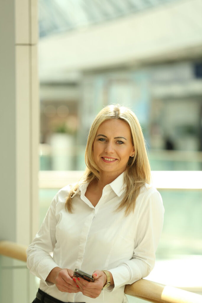Anna Suhajdova_CapitalMarkets - Tradeinfo.cz | Trader Magazine Professional woman with blonde hair in a white button-down shirt smiling while holding a smartphone, standing by a railing in a bright indoor atrium.