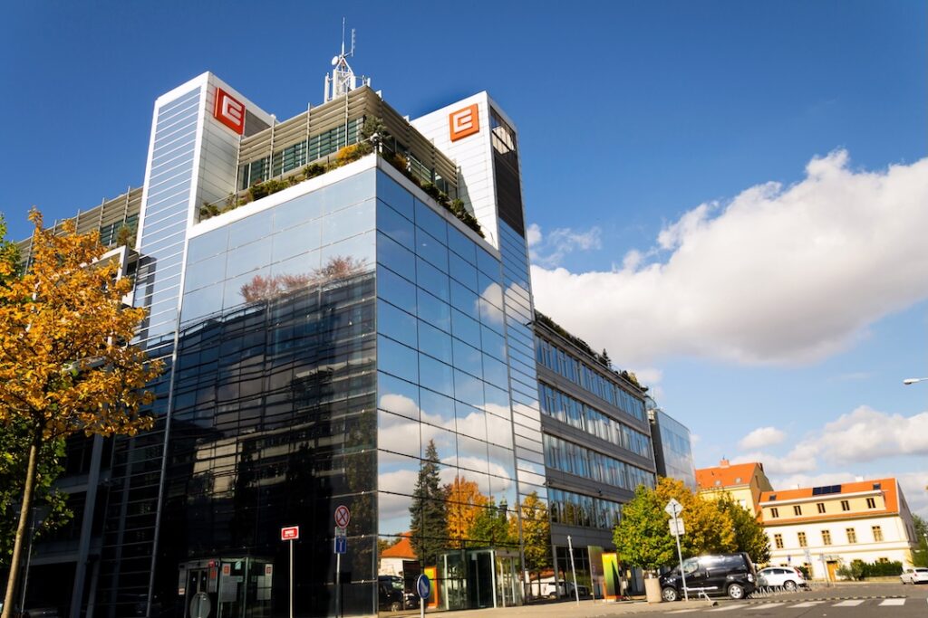 ČEZ - Tradeinfo.cz Modern glass office building with reflective facade, orange logos on upper corners, and autumn trees along the street.
