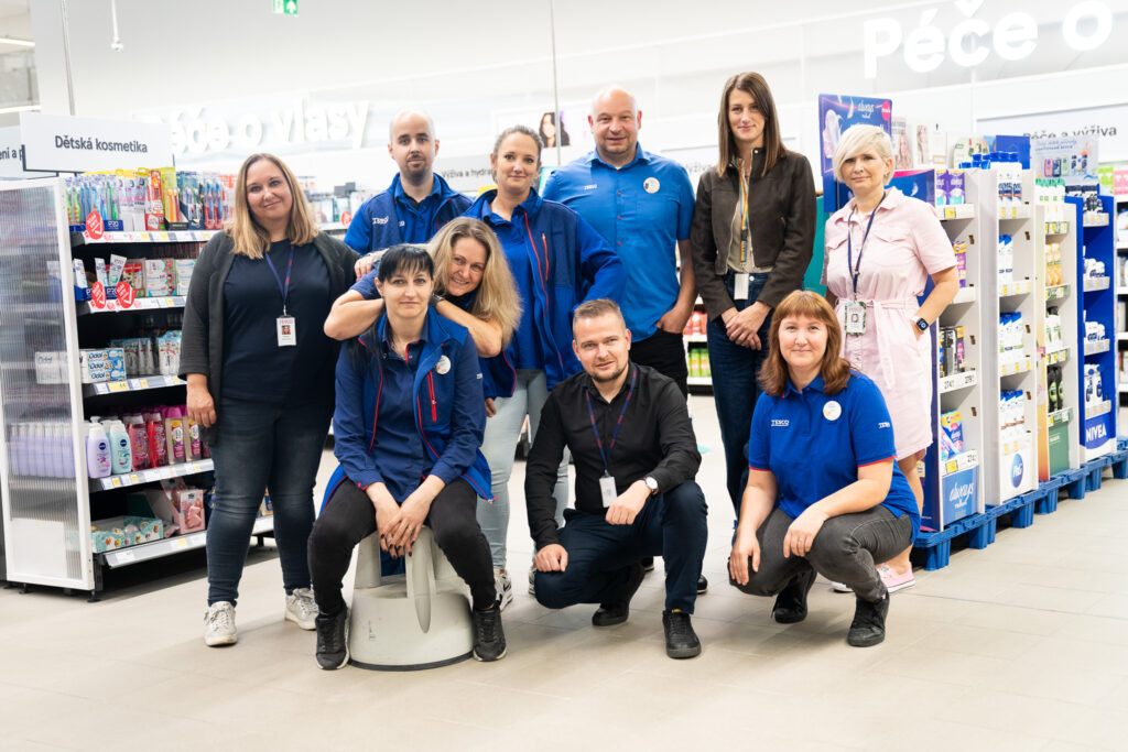 Péče o zaměstnance zůstává jednou z klíčových priorit společnosti - Tradeinfo.cz | Rozdílové smlouvy Group of store employees posing for a photo in a supermarket aisle, smiling at the camera.