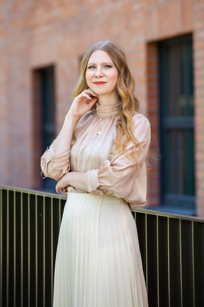 Olívia_Lacenova_photo - Tradeinfo.cz Portrait of a woman with long blonde hair in a beige blouse and cream pleated skirt, standing outdoors by a brick building with a railing, thoughtful pose.