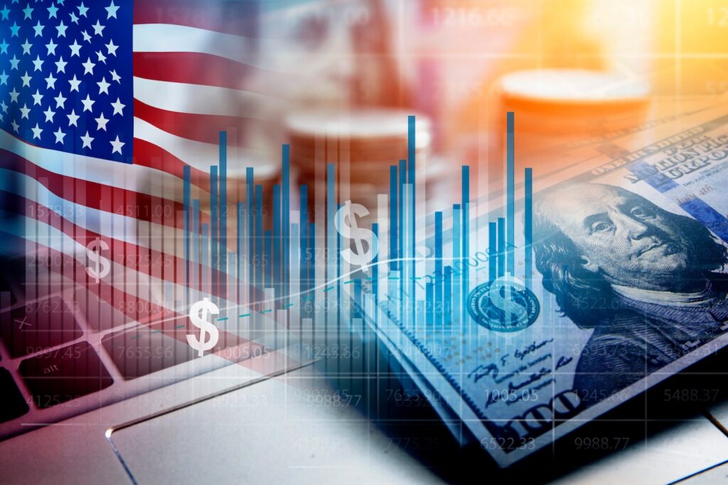 stocks USA5432 - Tradeinfo.cz American flag with overlaid financial bar charts, coins, and a hundred-dollar bill on a laptop, symbolizing U.S. finance and markets