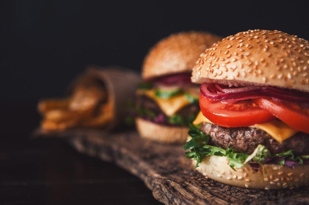Burger - Tradeinfo.cz Hamburger with sesame bun, lettuce, tomato, cheese, beef patty, and red onion on a wooden board, close-up focus on the front burger