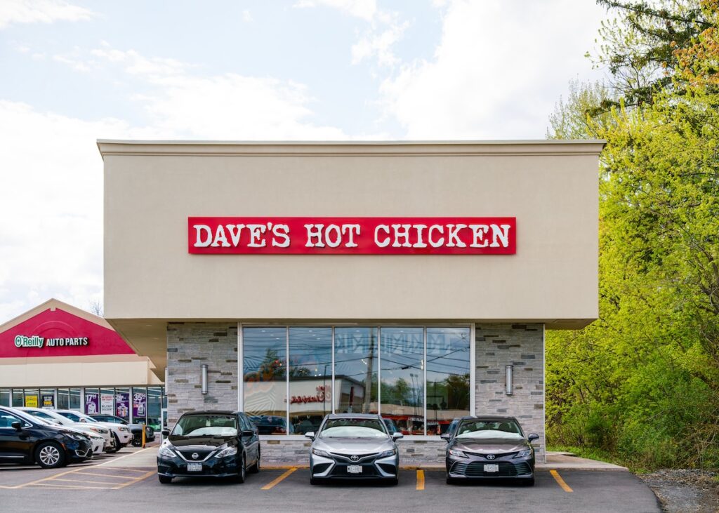 Daves Hot Chicken - Tradeinfo.cz Dave's Hot Chicken storefront with red sign and three cars parked in front during daytime contrastly lit by sun.