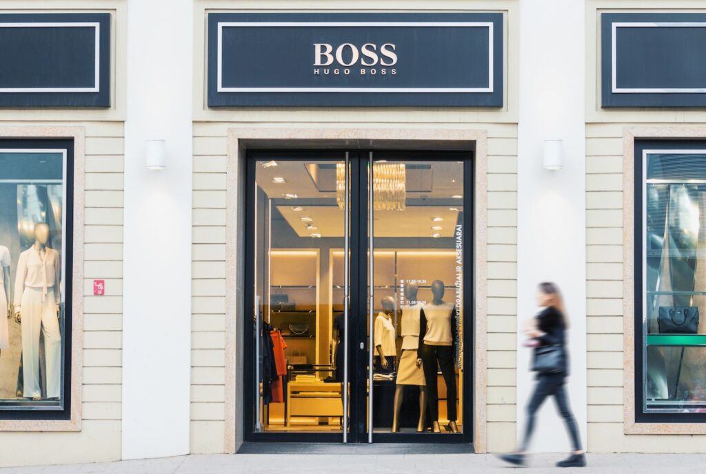 Hugo Boss - Tradeinfo.cz Hugo Boss storefront with glass double doors, mannequins inside, and a blurred passerby walking by a beige storefront.