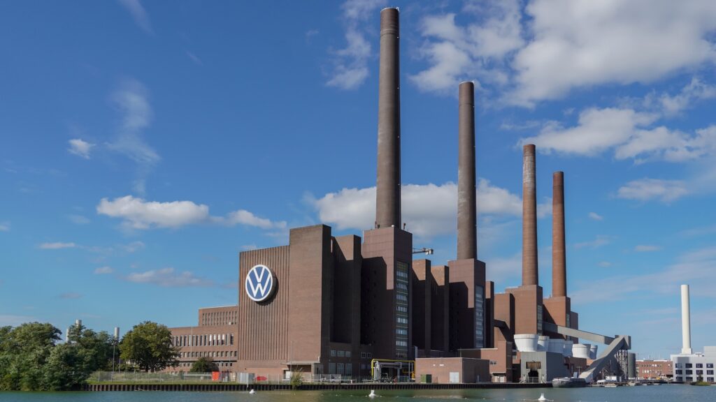 Volkswagen 6543 - Tradeinfo.cz Industrial brick factory by a river with four tall smokestacks and a VW logo on the facade, under a blue sky with clouds.