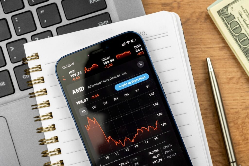 Akcie - Tradeinfo.cz Smartphone screen shows AMD stock chart with a red price line; notebook, pen, and a dollar bill in the background.