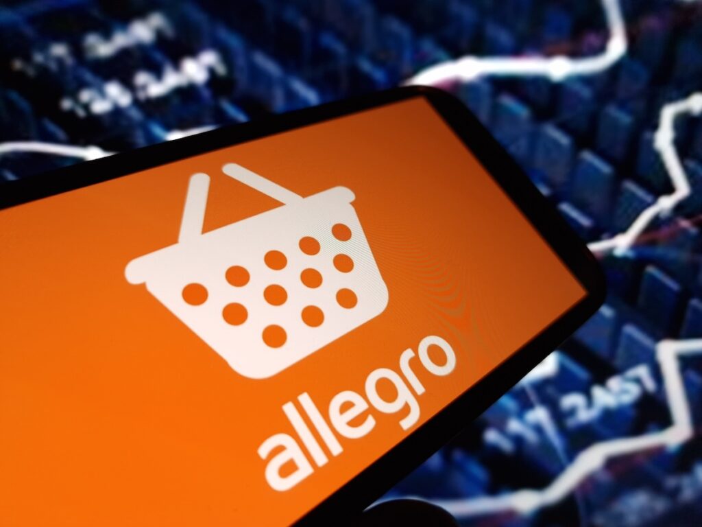 Allegro - Tradeinfo.cz Smartphone displaying the Allegro app logo on an orange screen with a blurred stock chart in the background short text description.