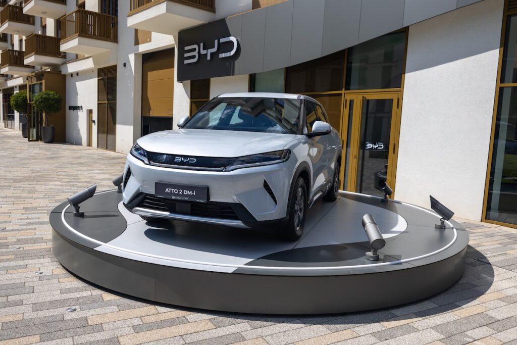 Automobil - Tradeinfo.cz White BYD Atto 2 DM-i SUV displayed on a circular showroom platform with spotlights and a BYD storefront in the background.