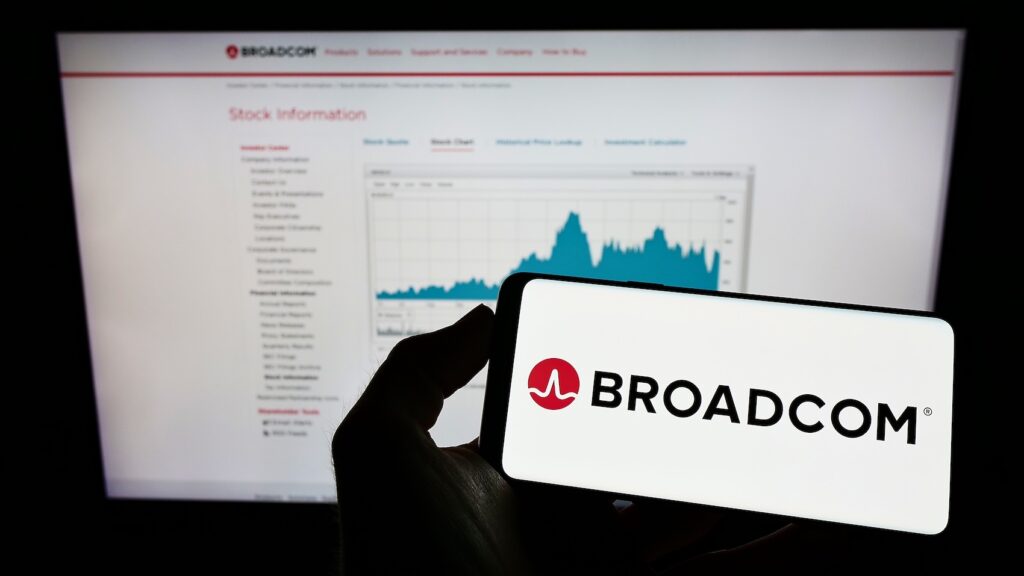 Broadcom - Tradeinfo.cz Close-up of a smartphone showing the Broadcom logo held in front of a computer screen displaying Broadcom stock information.