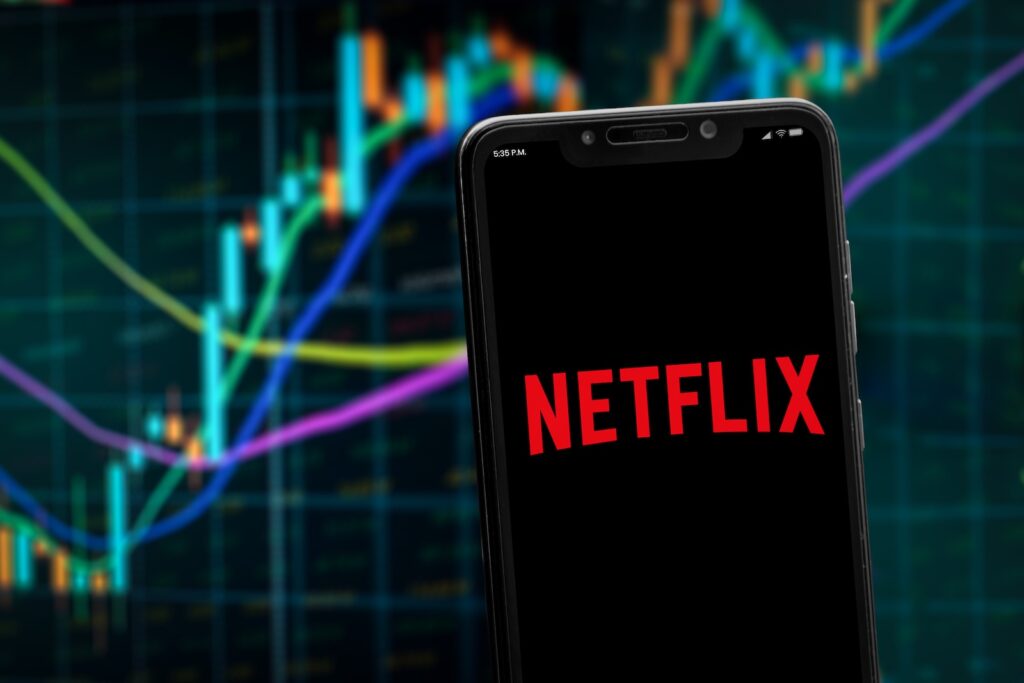NETFLIX - Tradeinfo.cz Smartphone displaying Netflix logo in red on a black screen with a blurred stock chart background behind it.