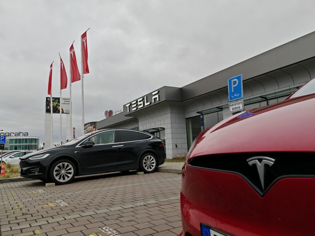 Tesla - Tradeinfo.cz Tesla showroom exterior with gray facade, red flagpoles, and multiple parked cars including a black sedan in the foreground.