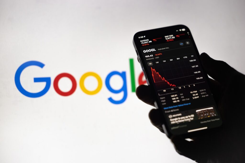GStock1111 - Tradeinfo.cz Phone screen shows Google stock chart with a red downward trend, held in front of a blurred Google logo in the background.