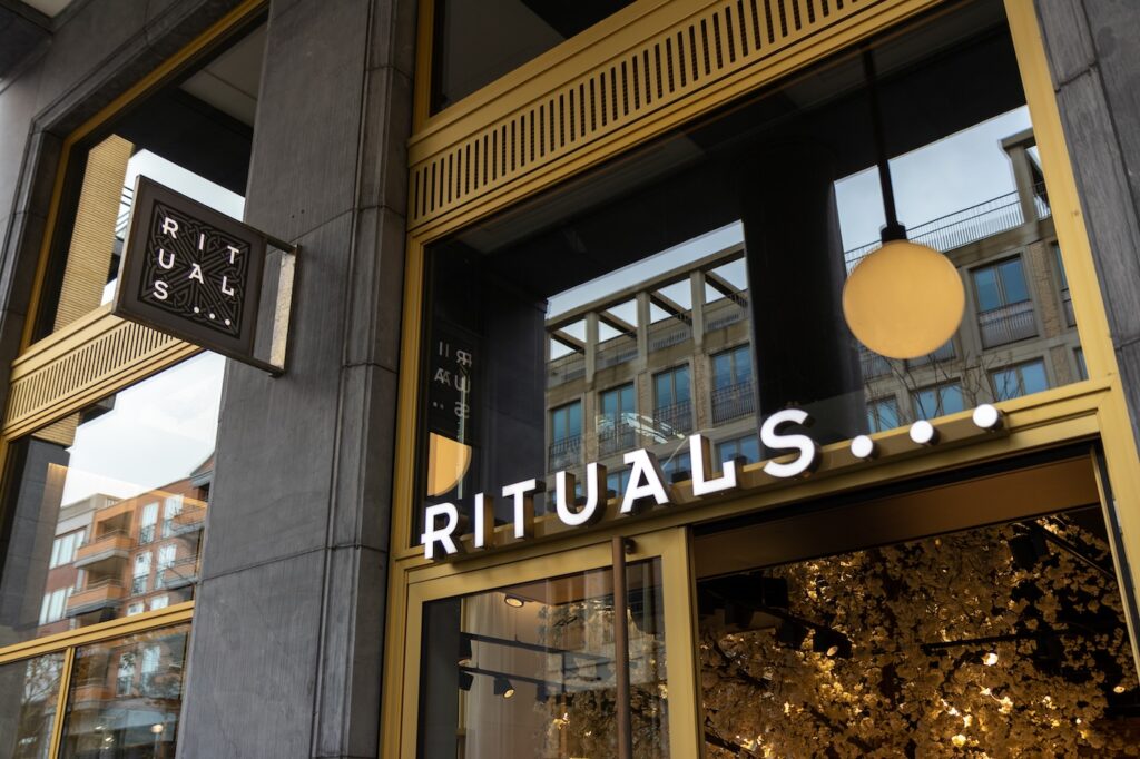 Rituals - Tradeinfo.cz Exterior of a Rituals store with a gold-framed glass storefront and a hanging sign displaying the brand name 'RITUALS' (ellipsis visible). The warm interior lighting and round yellow light fixture are visible inside.
