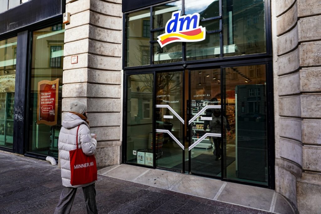 dm - Tradeinfo.cz Person in a white puffer jacket and beige hat stands outside a DM store with glass doors, carrying a red MINNER.HU tote bag.