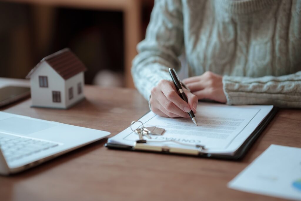 mortgage - hypotéka - Tradeinfo.cz Person signs a contract on a clipboard at a wooden desk, with a small model house nearby and a laptop in view